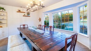 Comedor con una larga mesa de madera, seis sillas, una lámpara de araña y ventanas amplias con vistas a la vegetación.