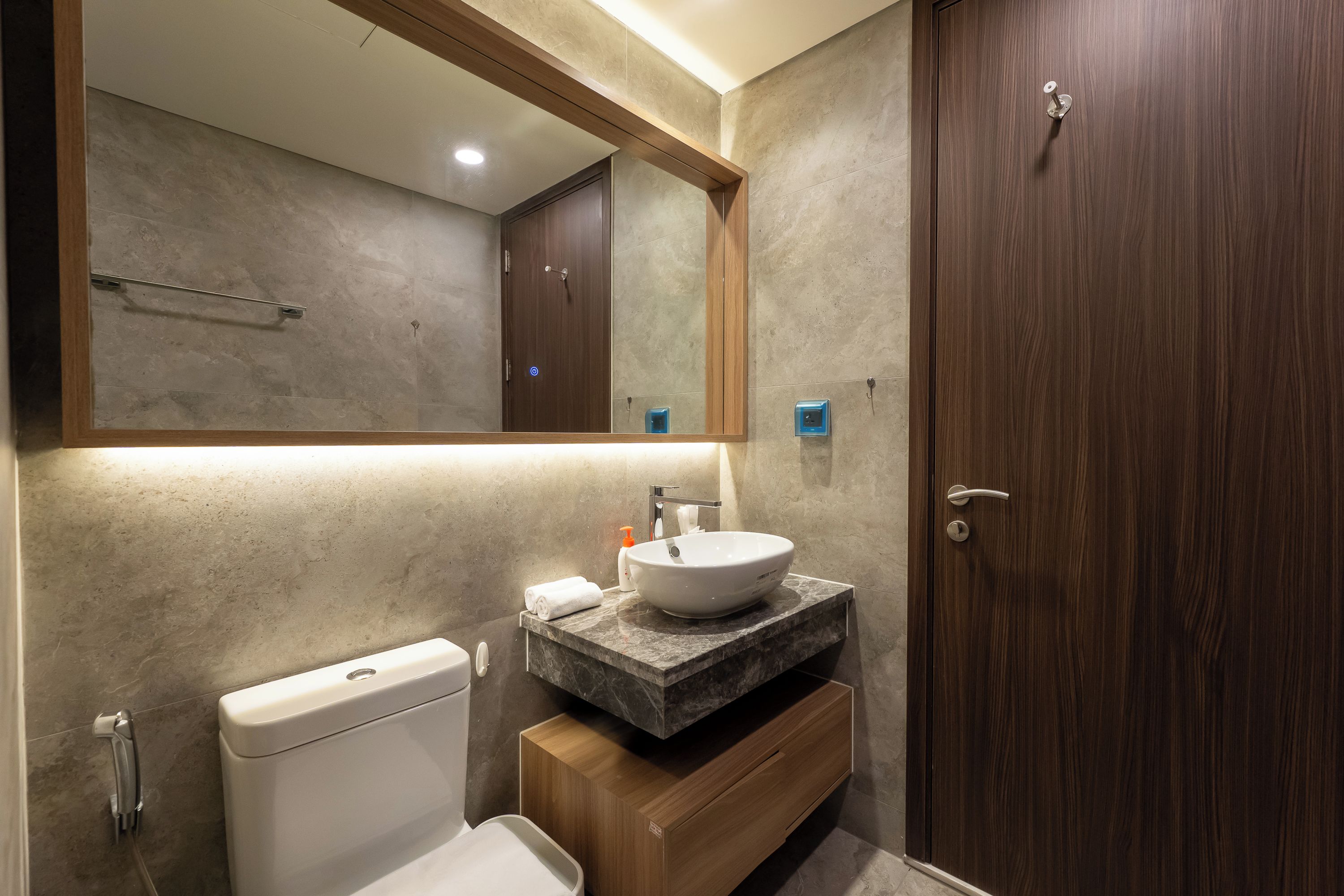 A modern bathroom with a marble countertop, a white sink, a wooden cabinet, a toilet, and a large mirror.
