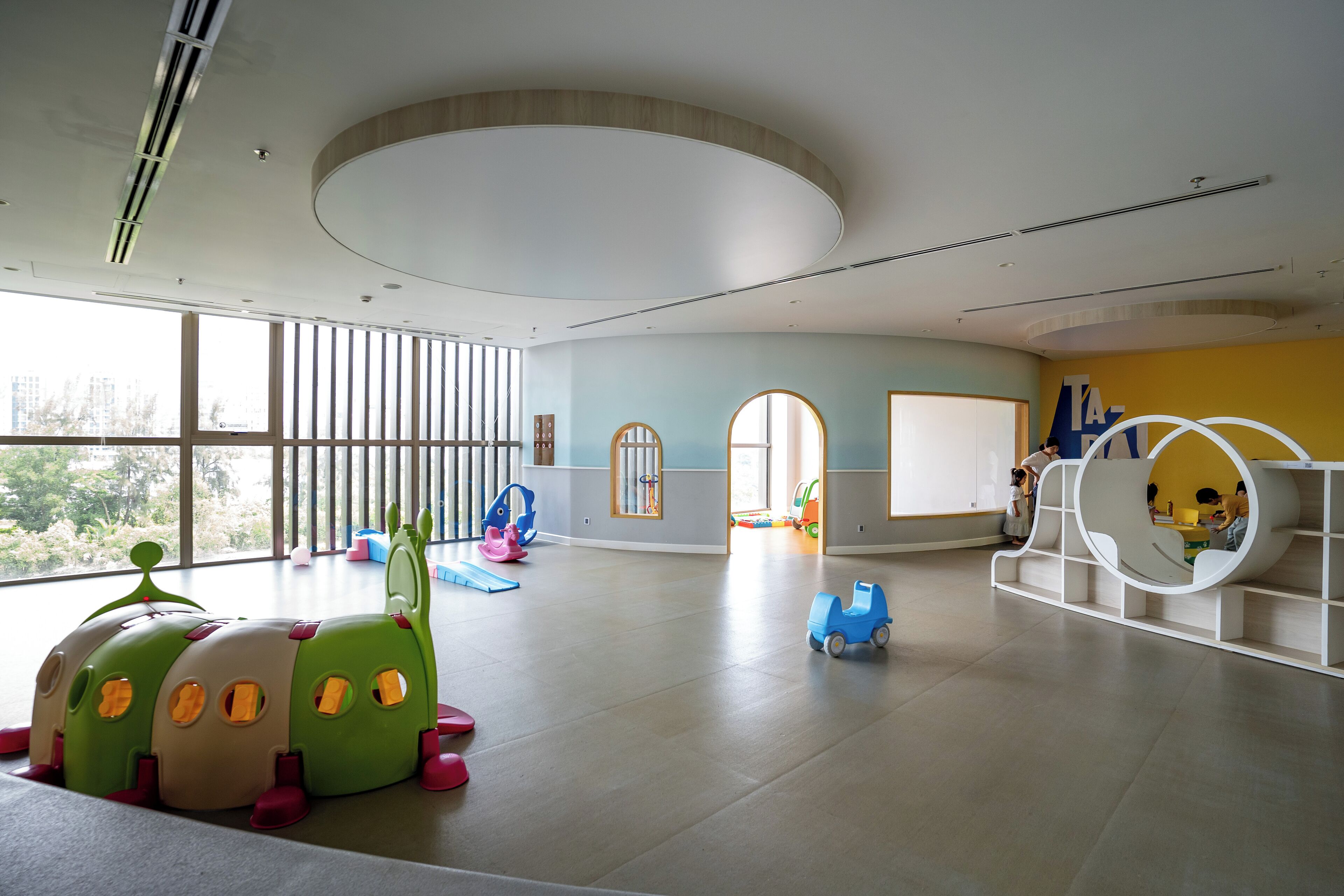 A spacious indoor play area with large windows, a circular ceiling design, and colorful play structures.