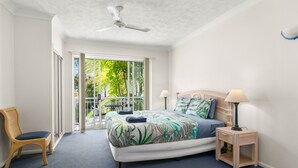 Quarto com cama, cadeira, mesa de cabeceira, ventilador de teto e vista para o verde através de uma porta deslizante.