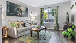 Sala de estar con sofá, mesa de centro y televisor. Hay dos ventanas con cortinas, una lámpara y una planta.