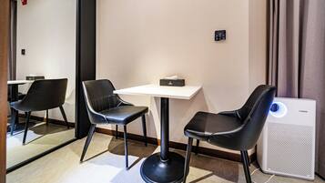 A hotel room with a white table, two black chairs, a small black box on the table, a wall-mounted control panel, and a white air purifier with a blue light.