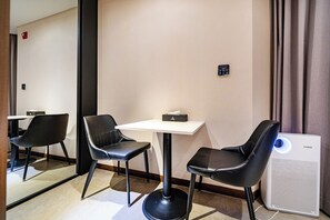 Une chambre d’hôtel avec une table blanche, deux chaises noires, une petite boîte noire posée sur la table, un panneau de commande fixé au mur et un purificateur d’air blanc doté d’une lumière bleue.
