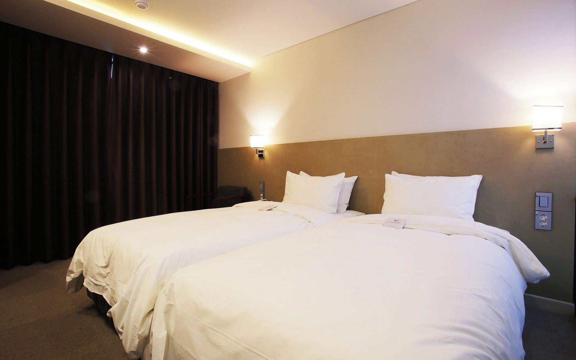 A hotel room with two beds, a dark curtain, a wall-mounted light, and a power outlet.