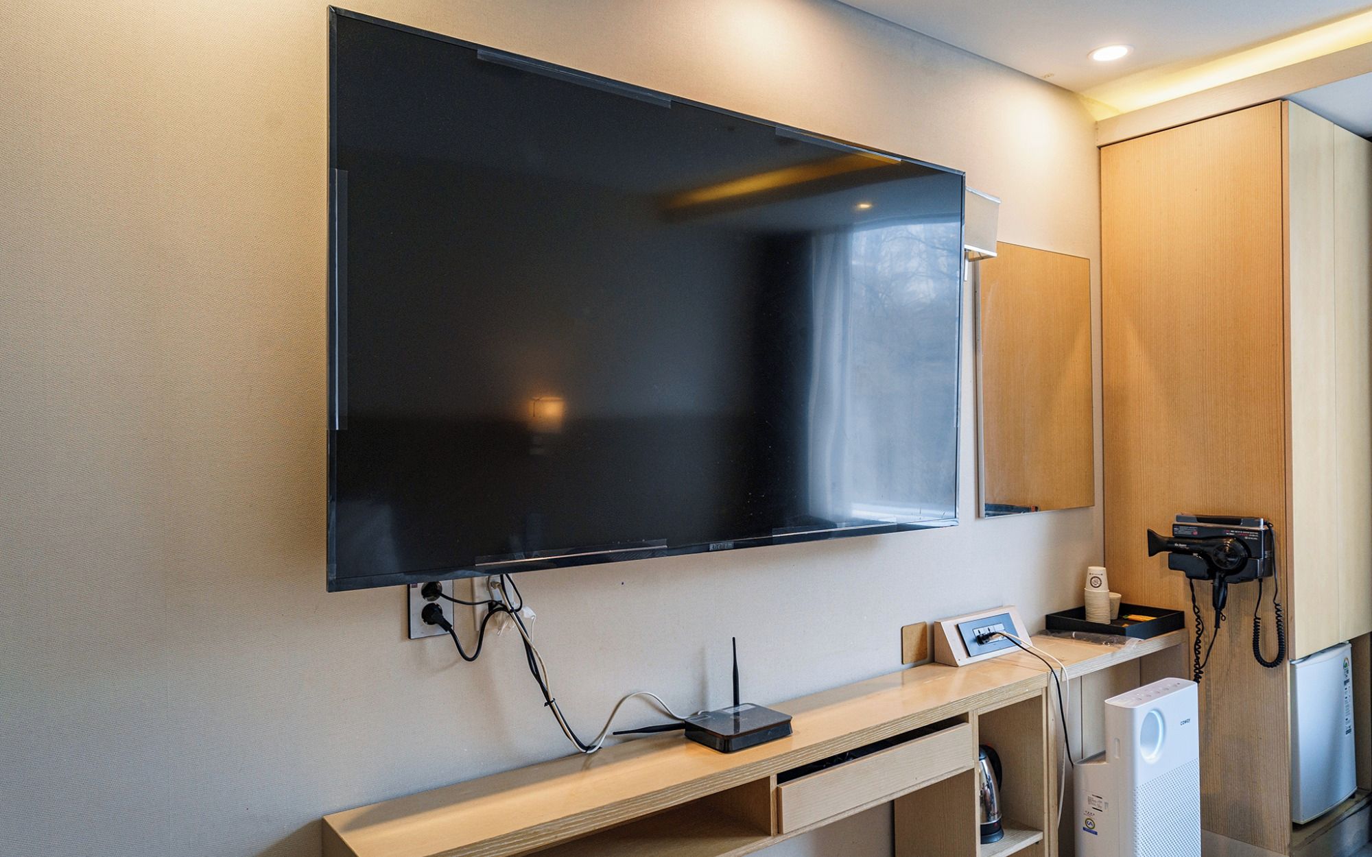 A modern hotel room with a large flat-screen TV mounted on the wall, a wooden console table with a coffee maker, and a compact refrigerator.