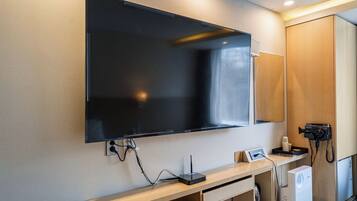 A modern hotel room with a large flat-screen TV mounted on the wall, a wooden console table with a coffee maker, and a compact refrigerator.