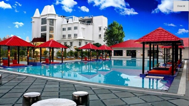 A swimming pool with red umbrellas and lounge chairs, surrounded by a building with multiple balconies. - Tiaraa Hotels & Resorts (Ramnagar)