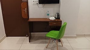 A small room with a wooden desk, a green chair, a television mounted on the wall, a water bottle, and a kettle. - CWR Stays (Noida)