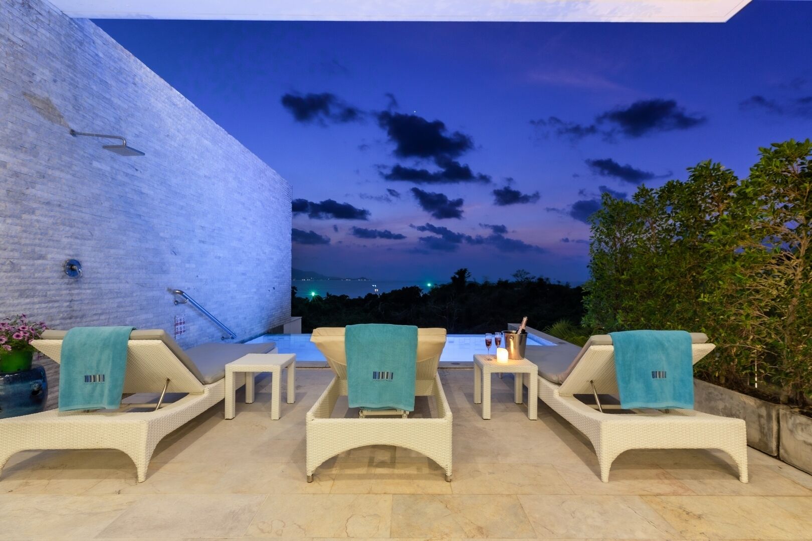 Un espace extérieur aménagé avec des fauteuils confortables et une piscine, le tout mis en valeur par un mur blanc et un ciel crépusculaire.