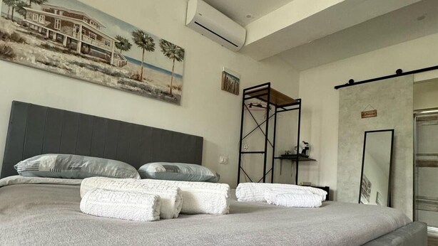 A large bed with grey linens and pillows, a headboard, an air conditioning unit, a painting of a house on the wall, and a metal rack with clothes. - Il Casello Country House  (Sciacca)