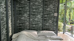 Un dormitorio con pared de diseño de piedra, cama con ropa de cama blanca y vista a la vegetación a través de un ventanal.
