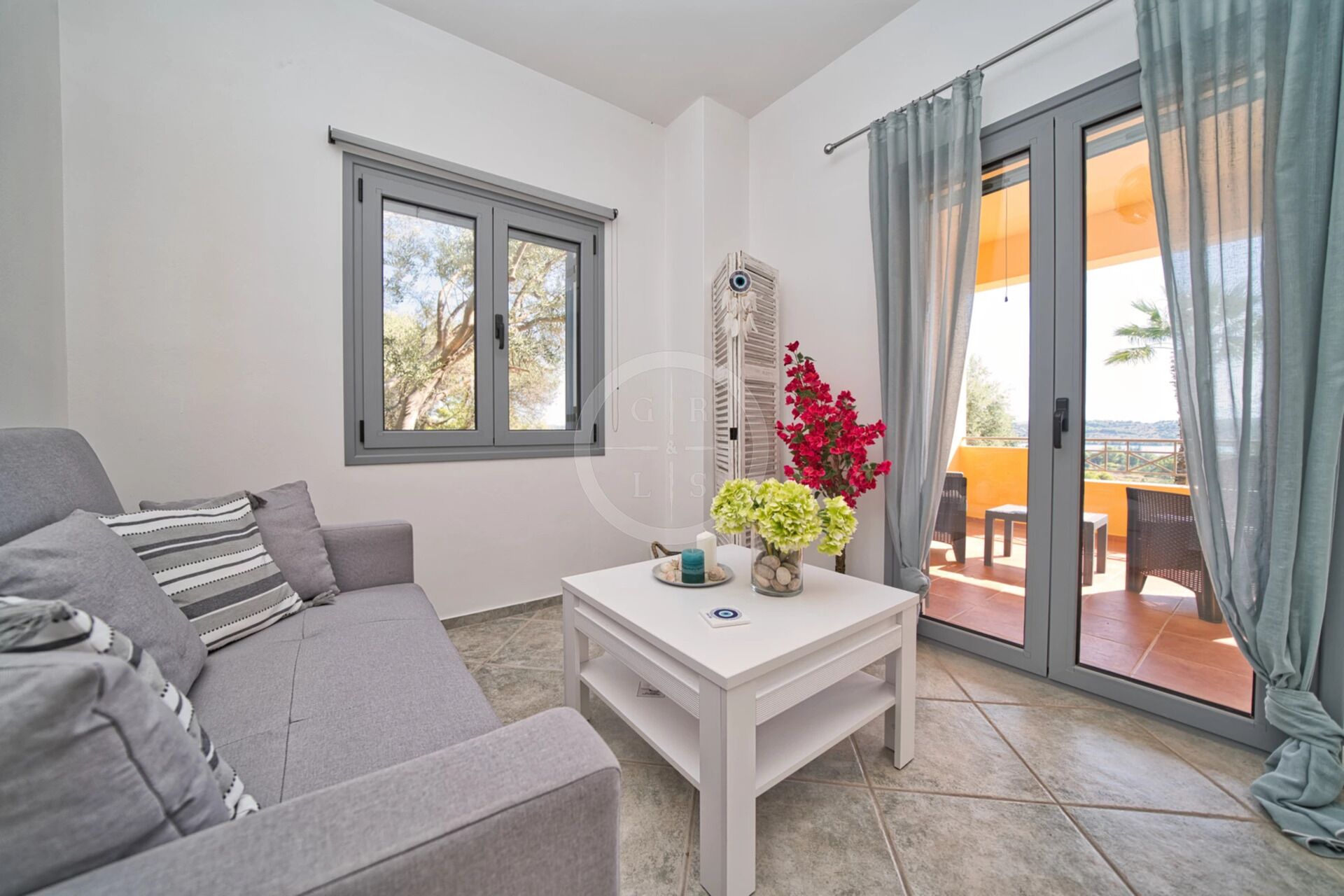 Ein modernes Wohnzimmer mit einem grauen Sofa, einem weißen Couchtisch und Blick durch eine Schiebetür auf eine Terrassen-Anlage.