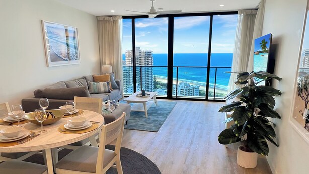 A modern living room with a dining area, large windows with a view of the ocean and buildings, a ceiling fan, and a potted plant.