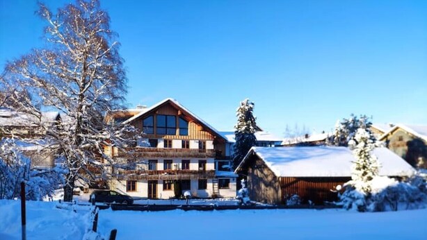 A multi-story wooden building with a snow-covered roof, surrounded by snow-covered trees and a clear blue sky. - Haus am Weißbach - Vacation in the Pfronten Valley (Pfronten)