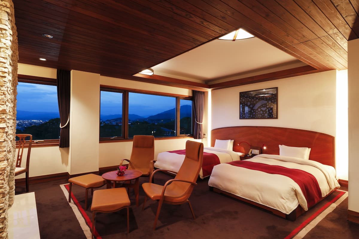 Quarto de hotel com duas camas, área de refeições com mesa e cadeiras, e vista para as montanhas através de grandes janelas.