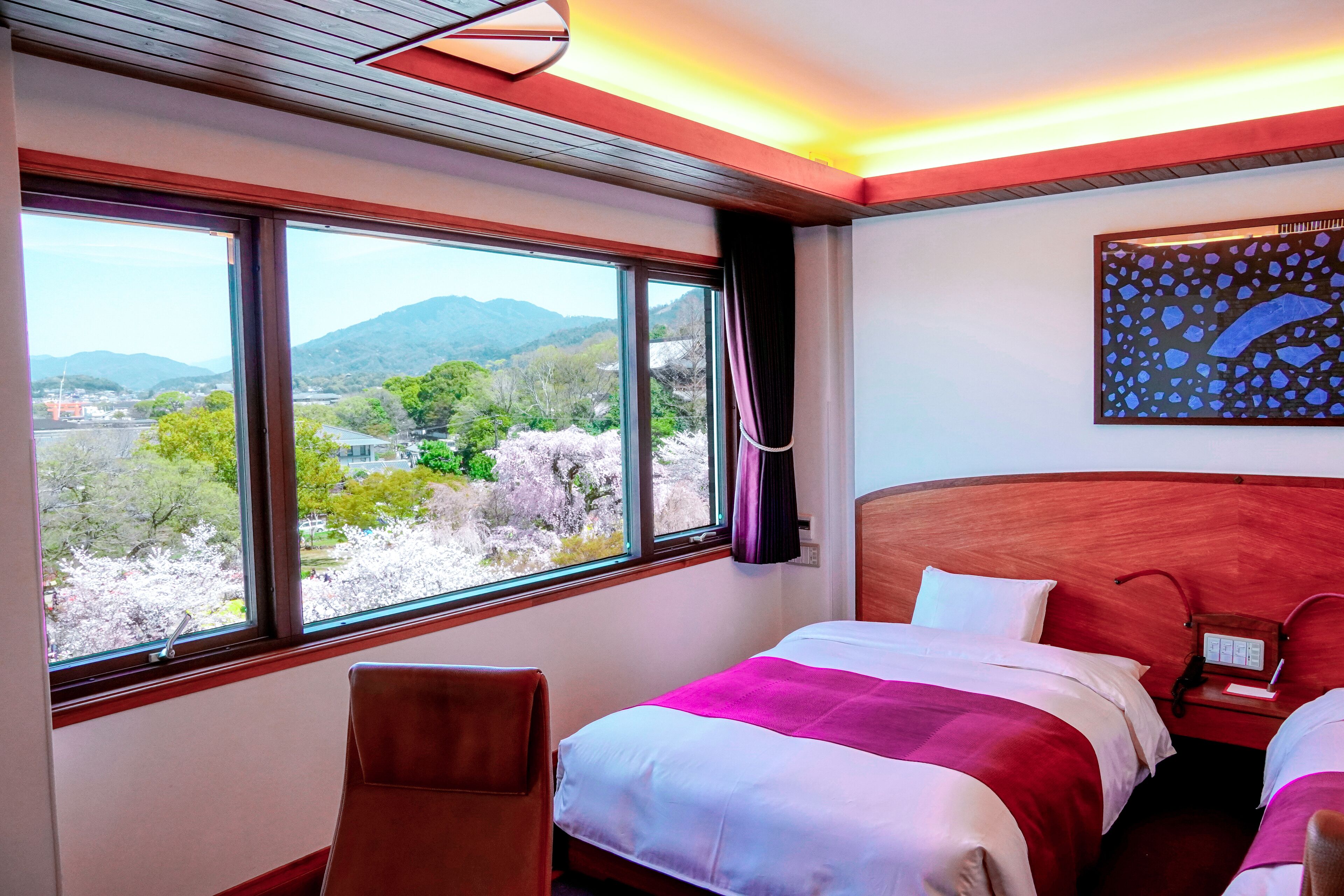 A hotel room with a large window offering a view of mountains and cherry blossoms. There is a bed with a red and white bedspread, a wooden headboard, and a nightstand with a lamp.