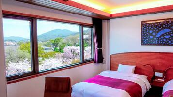 Une chambre d’hôtel dotée d’une grande fenêtre donnant sur un paysage de montagnes et de cerisiers en fleurs. On y trouve un lit recouvert d’une courtepointe rouge et blanche, une tête de lit en bois et une table de chevet avec une lampe.