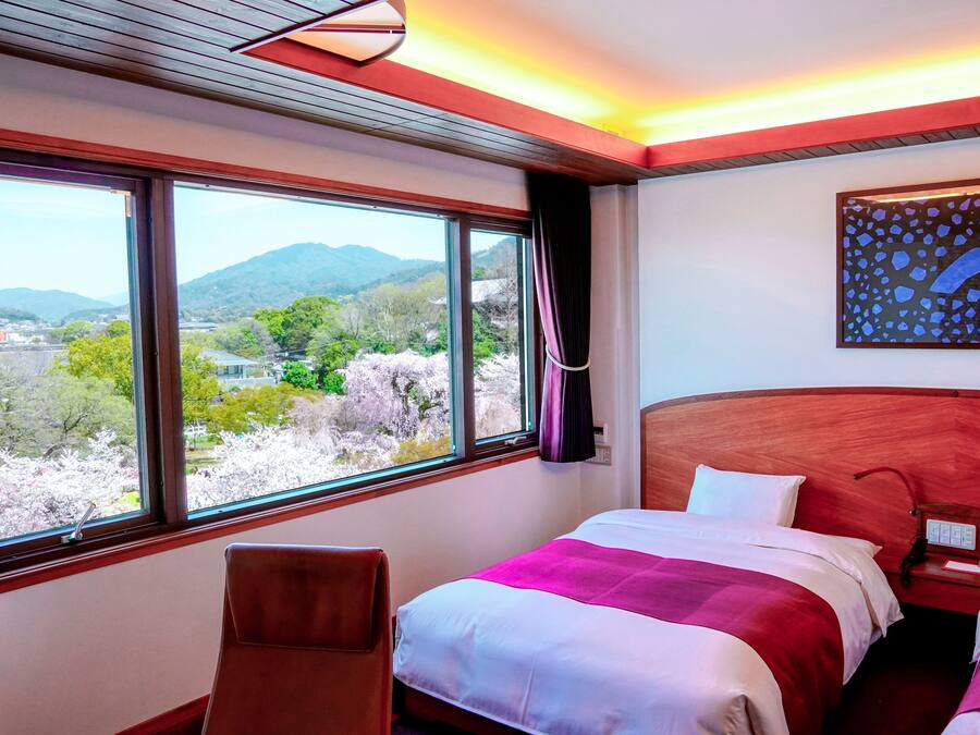 A hotel room with a large window offering a view of mountains and cherry blossoms. There is a bed with a red and white bedspread, a wooden headboard, and a nightstand with a lamp.