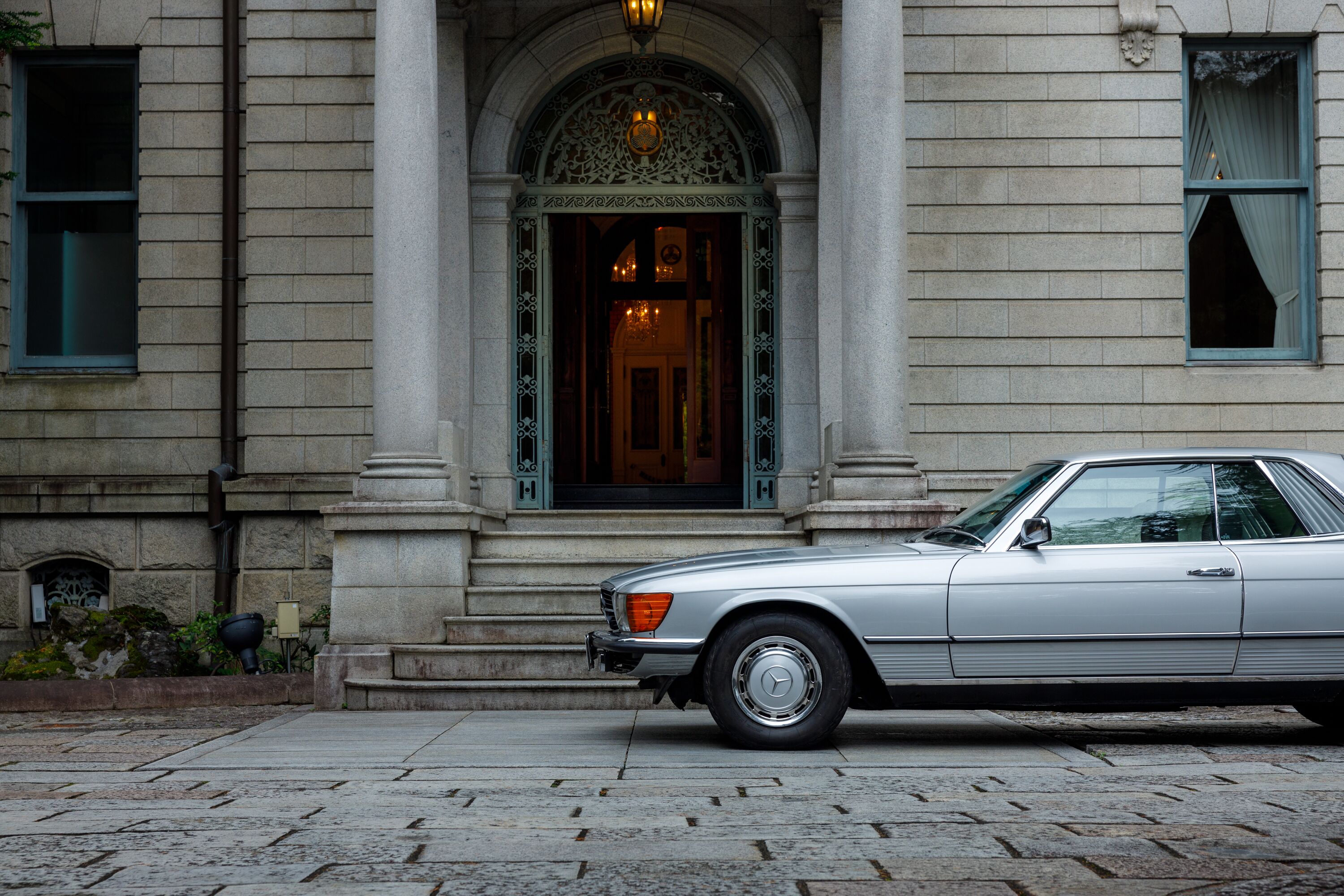 Une voiture argentée, modèle classique, est garée devant un immeuble à la façade en pierre, doté de grandes fenêtres et d’un portail d’entrée orné.