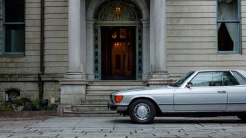 Une voiture argentée, modèle classique, est garée devant un immeuble à la façade en pierre, doté de grandes fenêtres et d’un portail d’entrée orné.