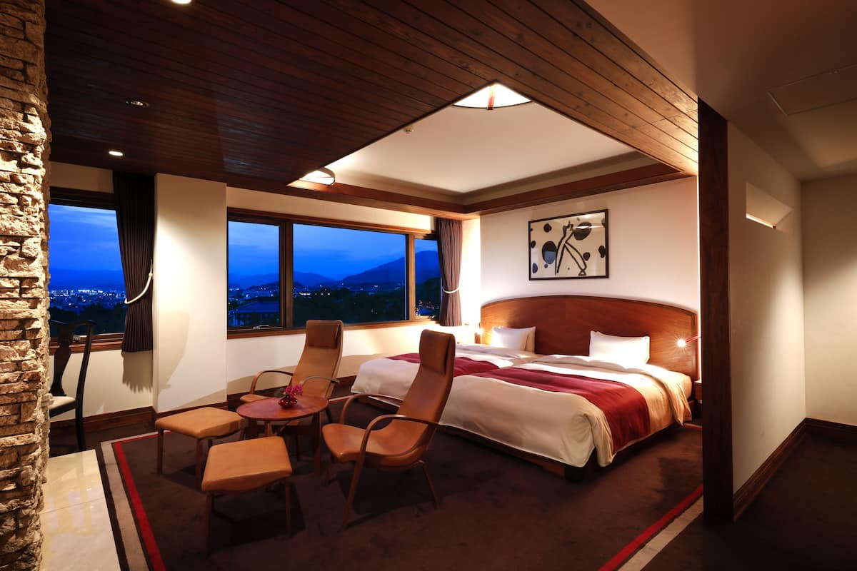 Quarto de hotel com uma cama grande, duas cadeiras, uma mesinha e vista para as montanhas.