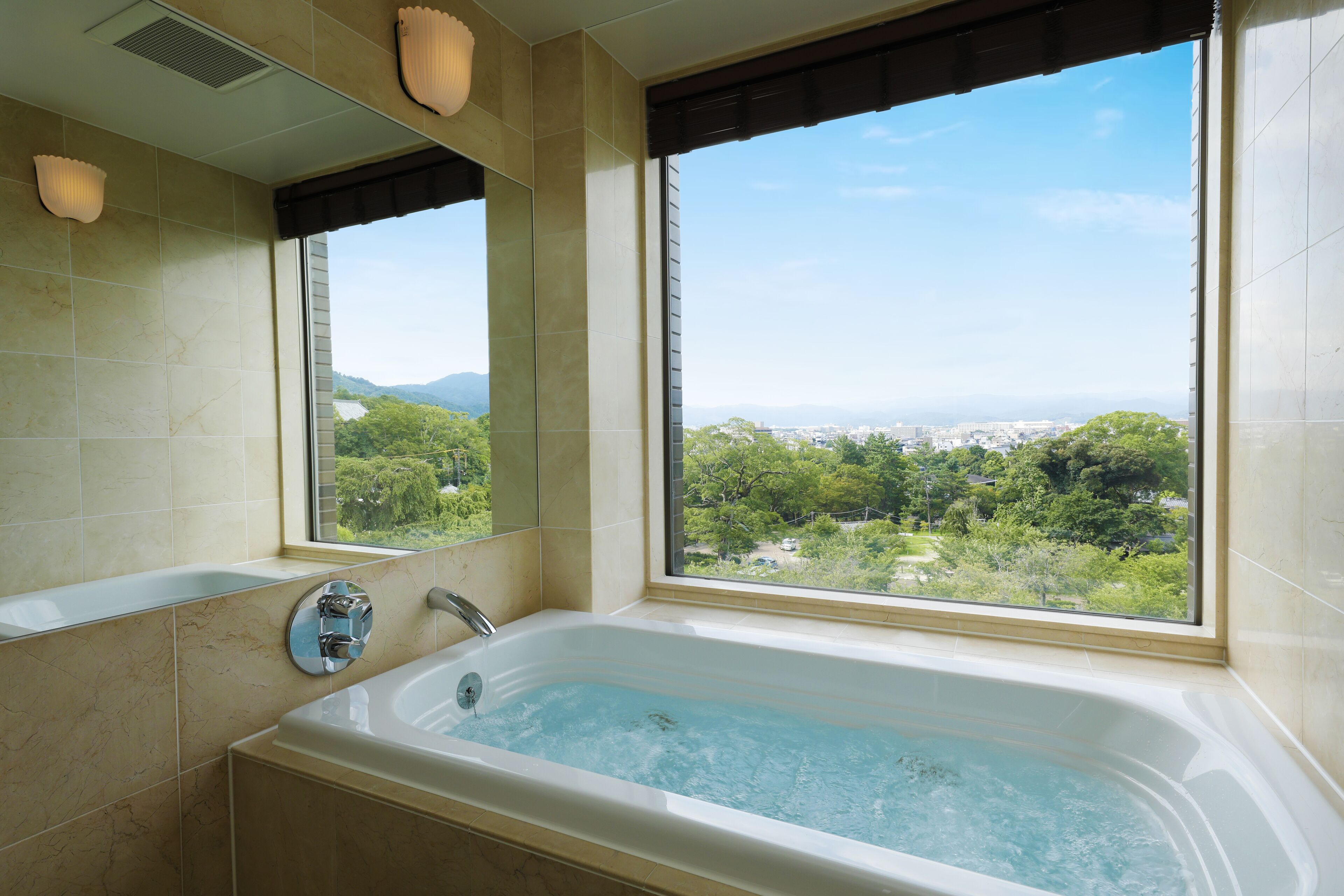 Un baño con bañera, ventana con vistas a zonas verdes y edificios, y un espejo.