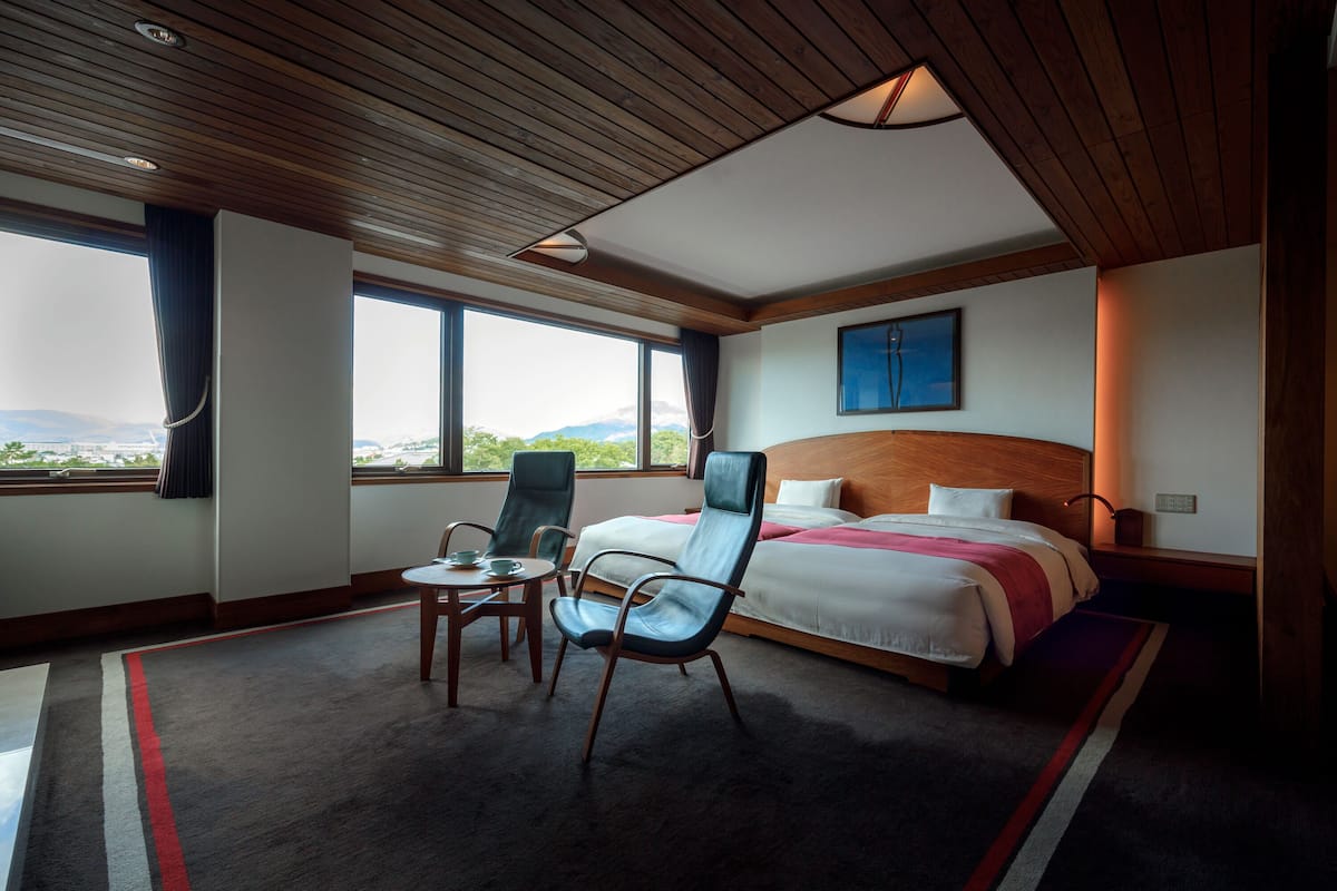 Quarto de hotel com uma cama grande, duas cadeiras, uma mesa e vista para as montanhas por grandes janelas.