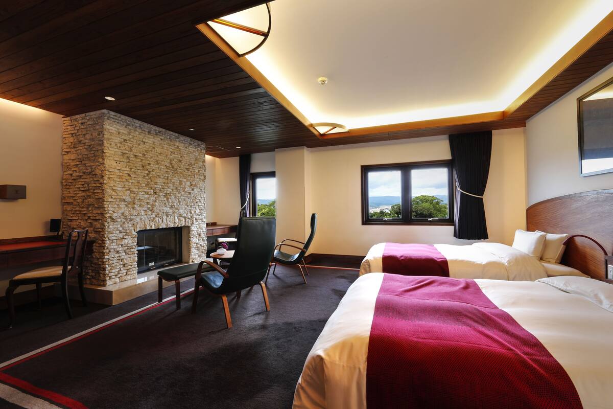 Quarto de hotel com duas camas, uma lareira de pedra, uma escrivaninha com cadeira e janelas amplas com vista.