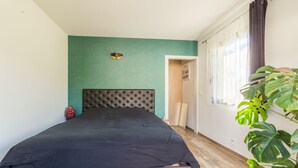Ett sovrum med en stoppad sänggavel, en säng med ett mörkt överkast, ett trägolv, en grön accentvägg, ett fönster med gardiner och en krukväxt.