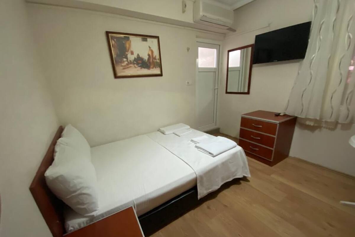 Une chambre avec un lit simple, un plancher en bois, une table de chevet, une armoire, un tableau au mur et une fenêtre avec des rideaux.