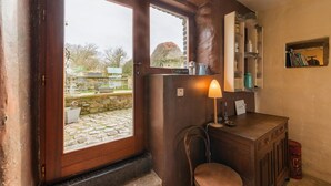 A rustic room with a wooden door, a small table with a lamp, a chair, and a shelf with kitchen items.