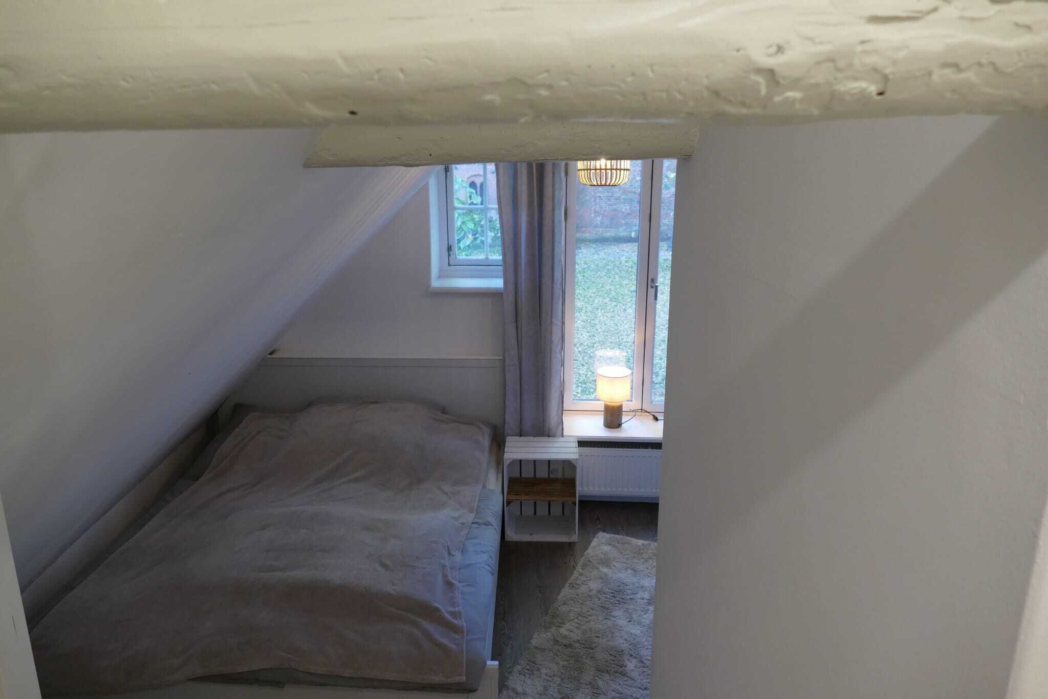 A small, narrow room with a sloped ceiling, a single bed, a radiator, and a window with a lamp.