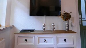 Ett rum med ett träpanelat tak, en väggmonterad platt-TV, ett vitt skåp med lådor och en vas med blommor på väggen.