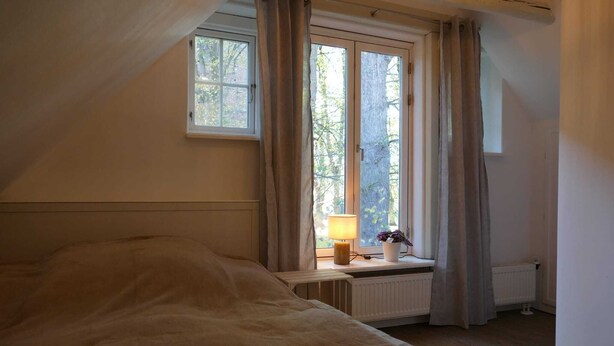 Ett sovrum med en säng, ett fönster med gardiner, ett element och ett litet bord med en lampa och en växt.
