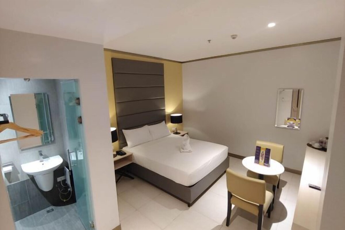 A modern hotel room with a large bed, bedside table, lamp, and a small round table with a chair.