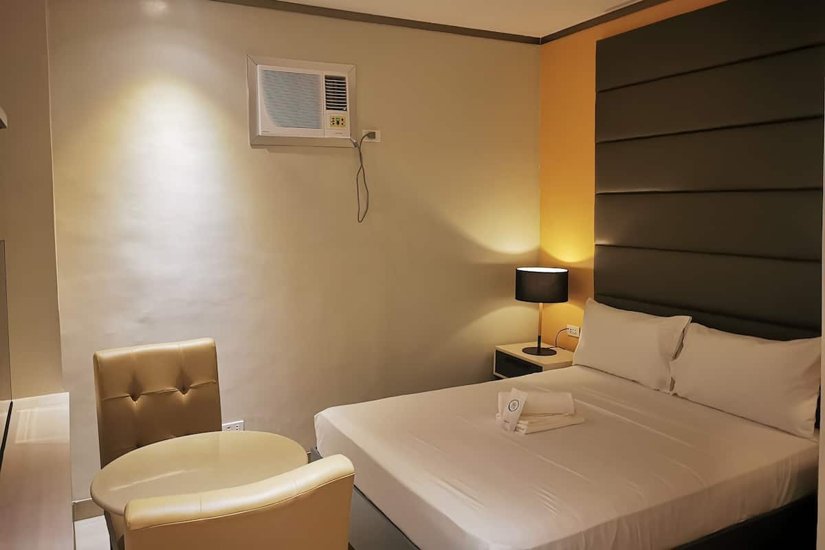 A hotel room with a bed, a chair, a nightstand, a lamp, and an air conditioning unit.