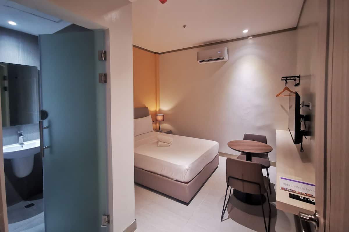 A modern hotel room with a bed, bedside table, chair, and a small table with a chair. There is a mirror, a wall-mounted TV, and an air conditioning unit.