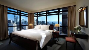 Habitación de hotel con una cama grande, dos lámparas de noche, un escritorio con teléfono, una silla, un espejo y vistas a un paisaje urbano a través de ventanas amplias.