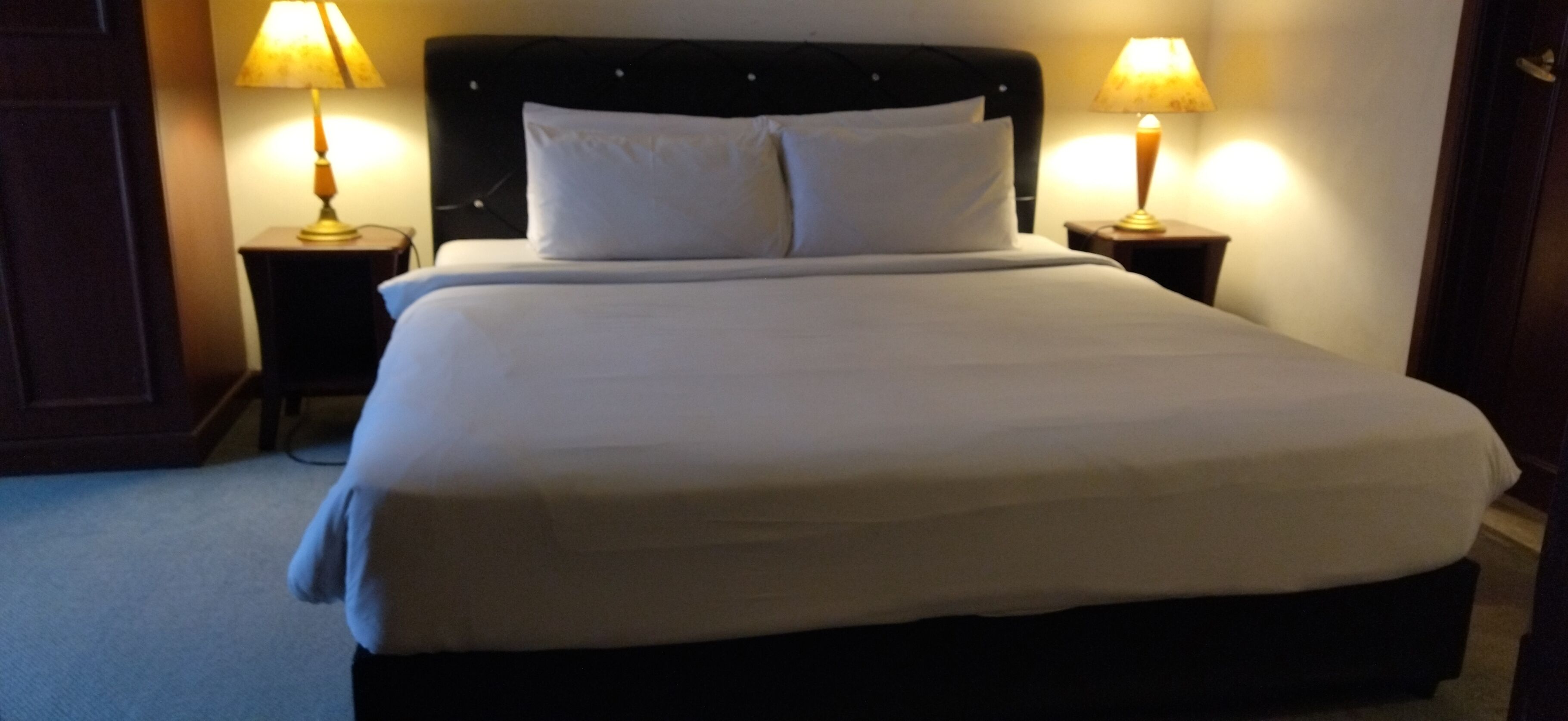 A neatly made bed with white linens, two bedside lamps, and a dark headboard.