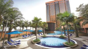 Un área de piscina hotelera con sillones de descanso y sombrillas, rodeada de palmeras y un edificio alto.