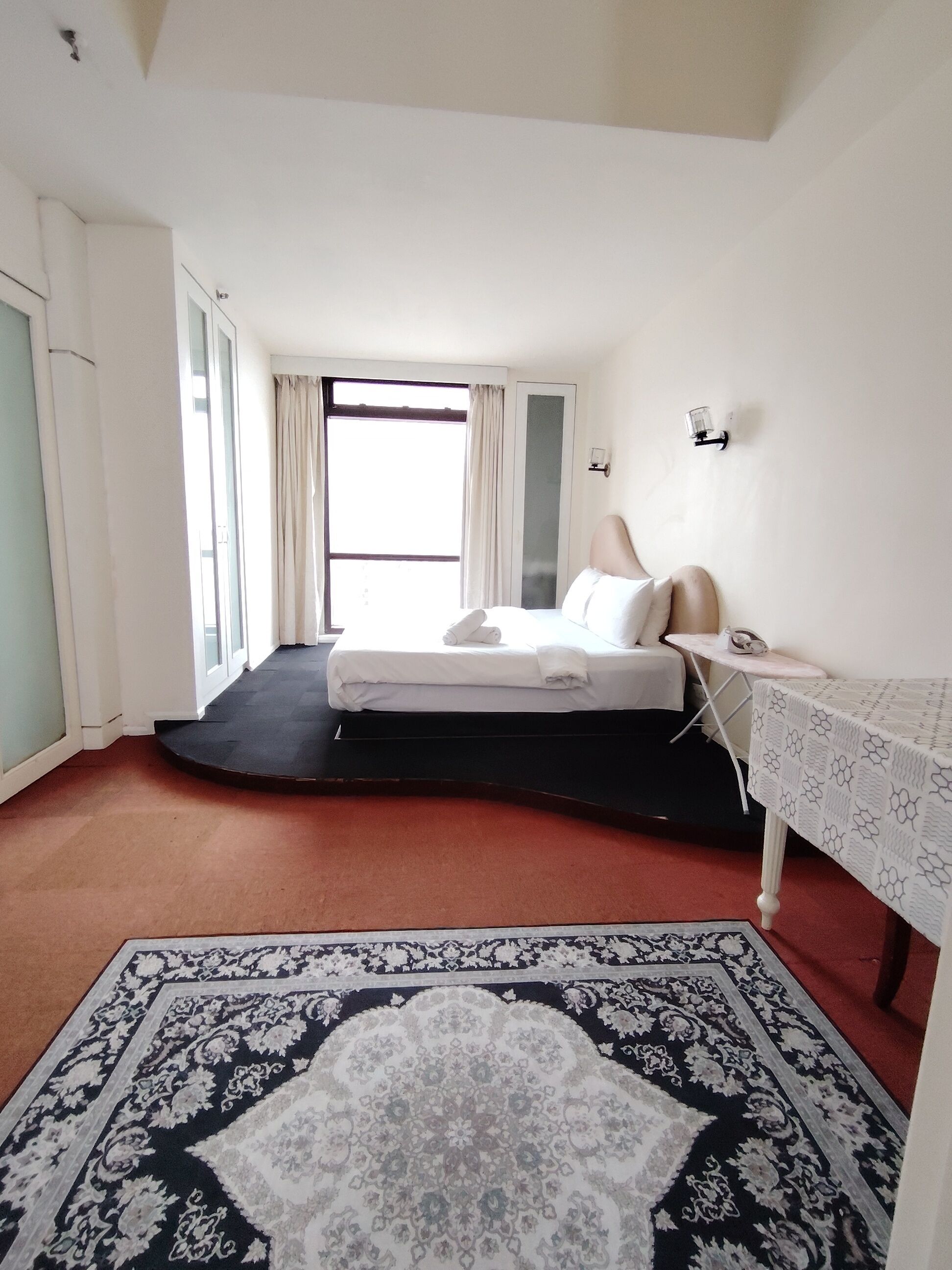 A hotel room with a bed, a small table, a nightstand, a large window with curtains, and a patterned rug.