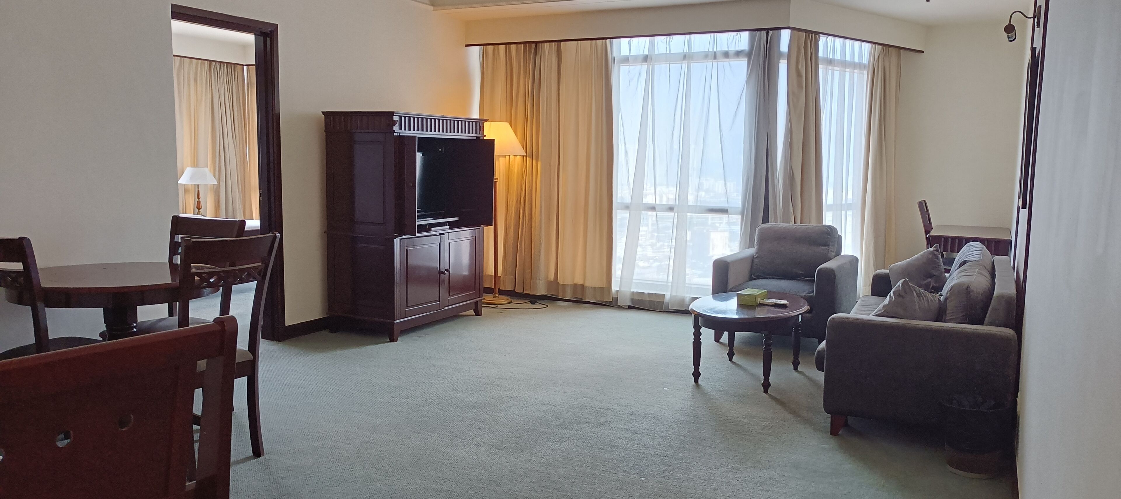 Une chambre d’hôtel comprenant une table à manger, deux fauteuils, un canapé, une télévision et une grande fenêtre avec des rideaux.