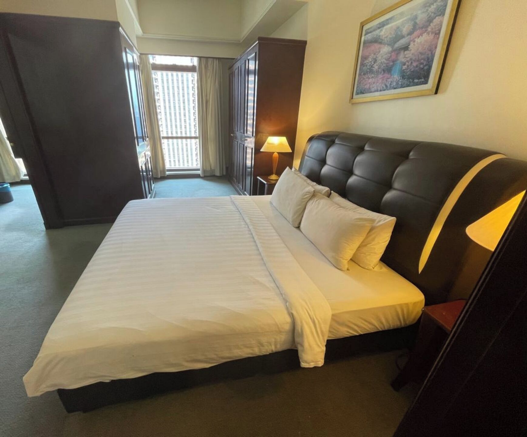A hotel room with a large bed, a nightstand with a lamp, a wardrobe, and a view of the outside through a window.