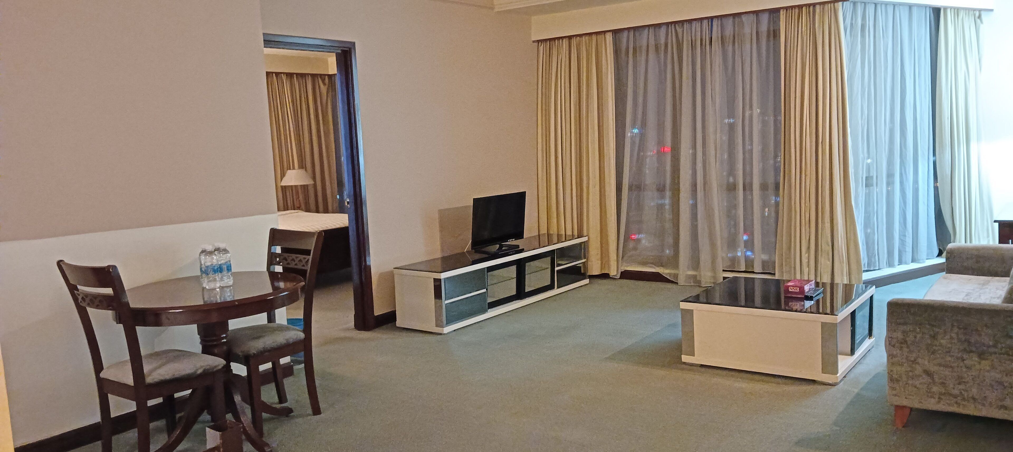 A hotel room with a dining table, chairs, a TV, a sofa, and a large window with curtains.