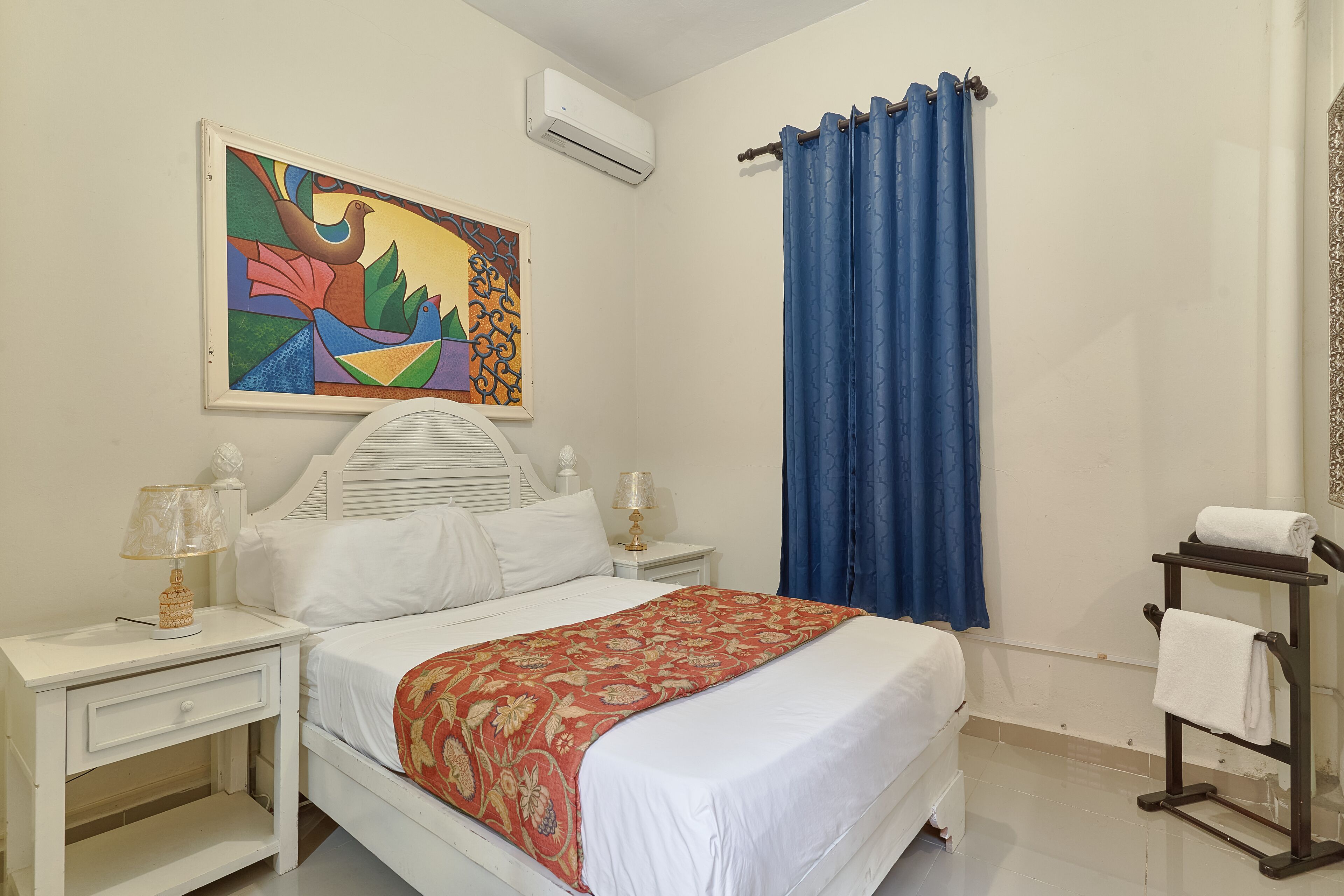 Une chambre à coucher comprenant un lit, deux tables de chevet, un climatiseur fixé au mur, un rideau bleu et un porte-serviettes.