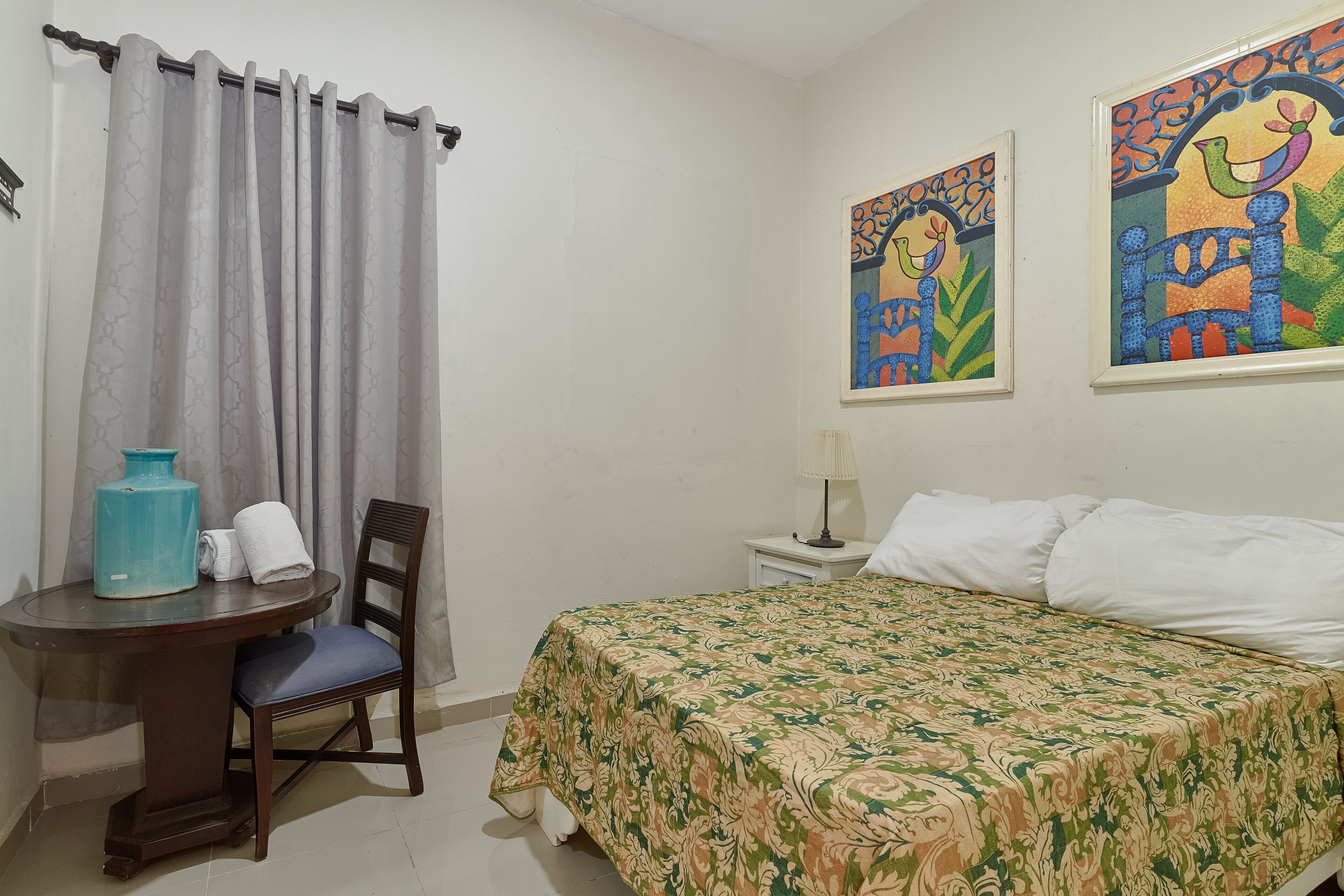 A hotel room with a bed, a chair, a small table with a vase and towels, and two paintings on the wall.