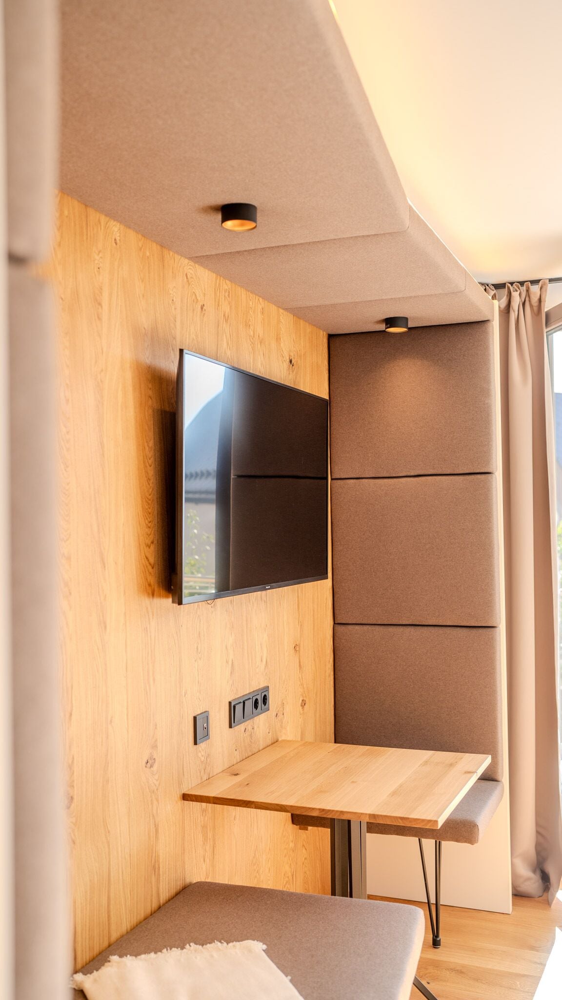 A modern RV interior with a flat-screen TV mounted on a wooden wall, a table, and a cushioned bench.