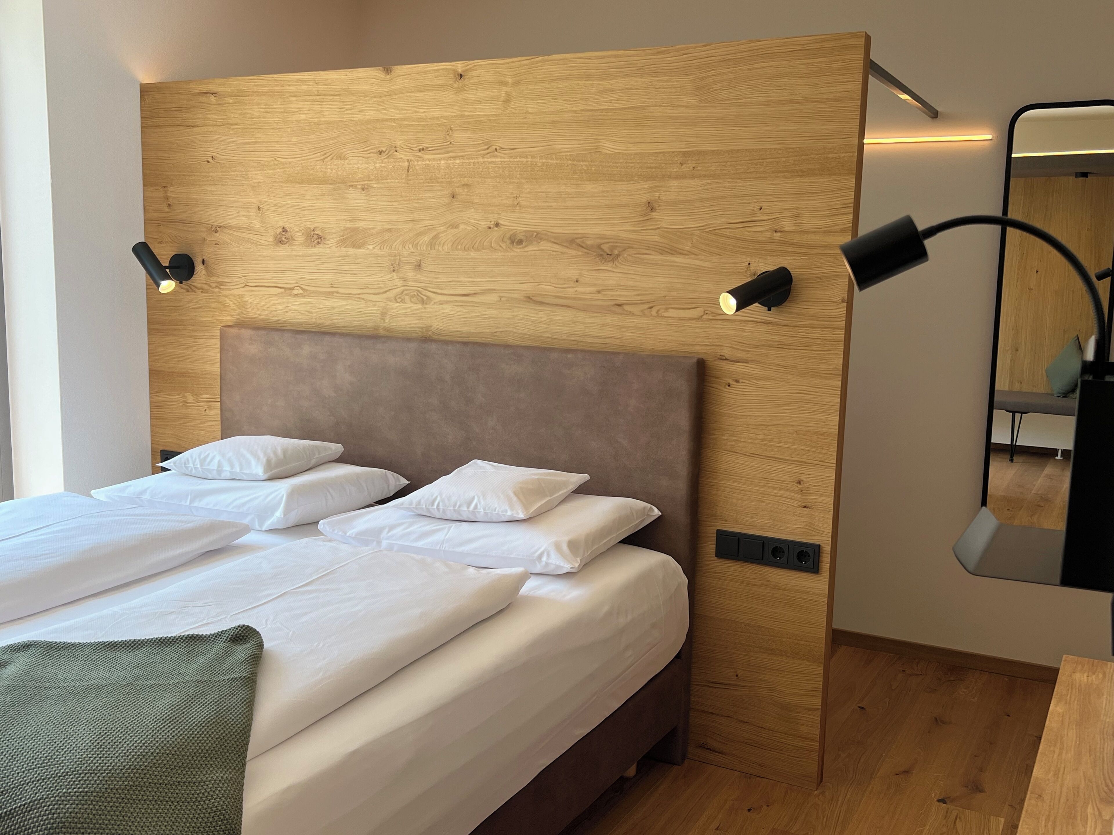 Un dormitorio moderno con cabecera de madera, cama con ropa blanca, dos lámparas de noche y un espejo en la pared.