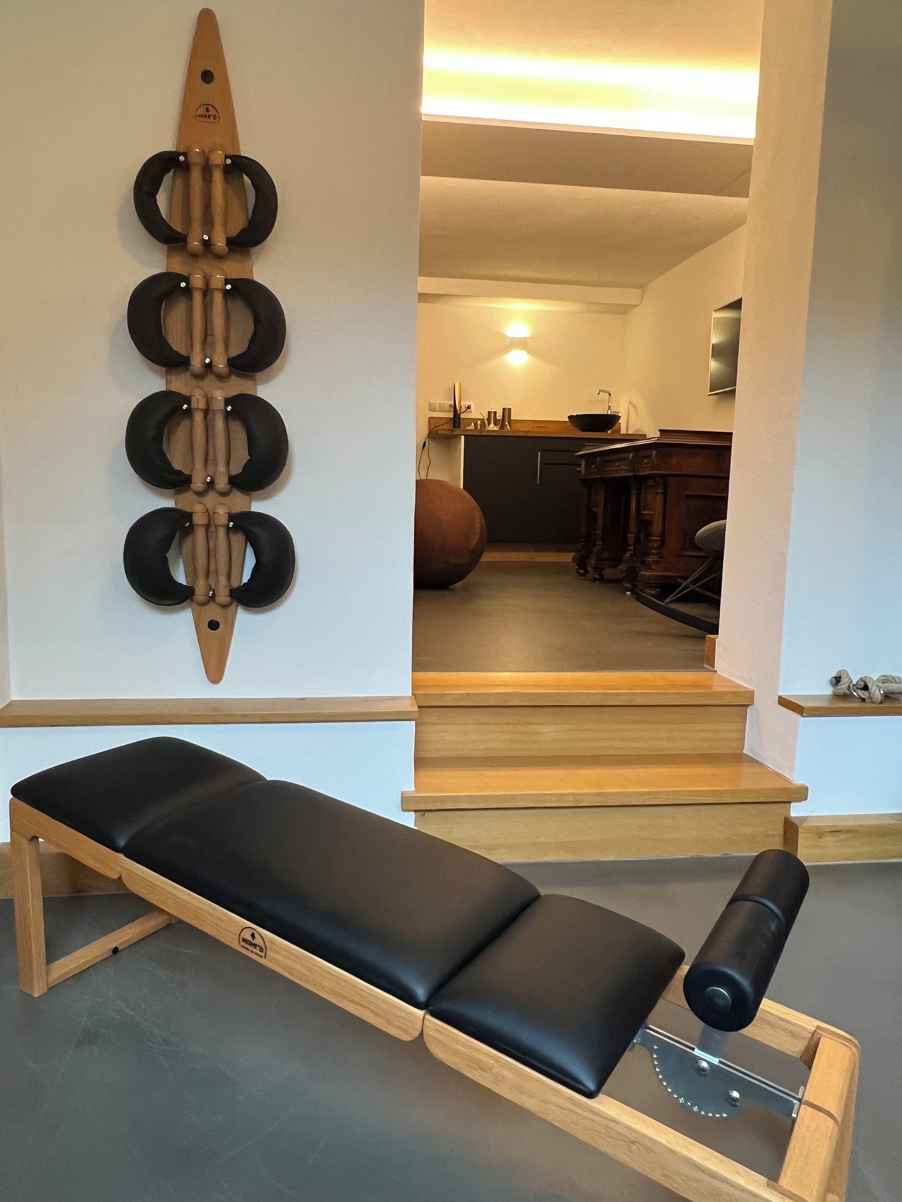 A room with a wooden bench, a wall-mounted exercise device, a kitchen area, and a staircase.