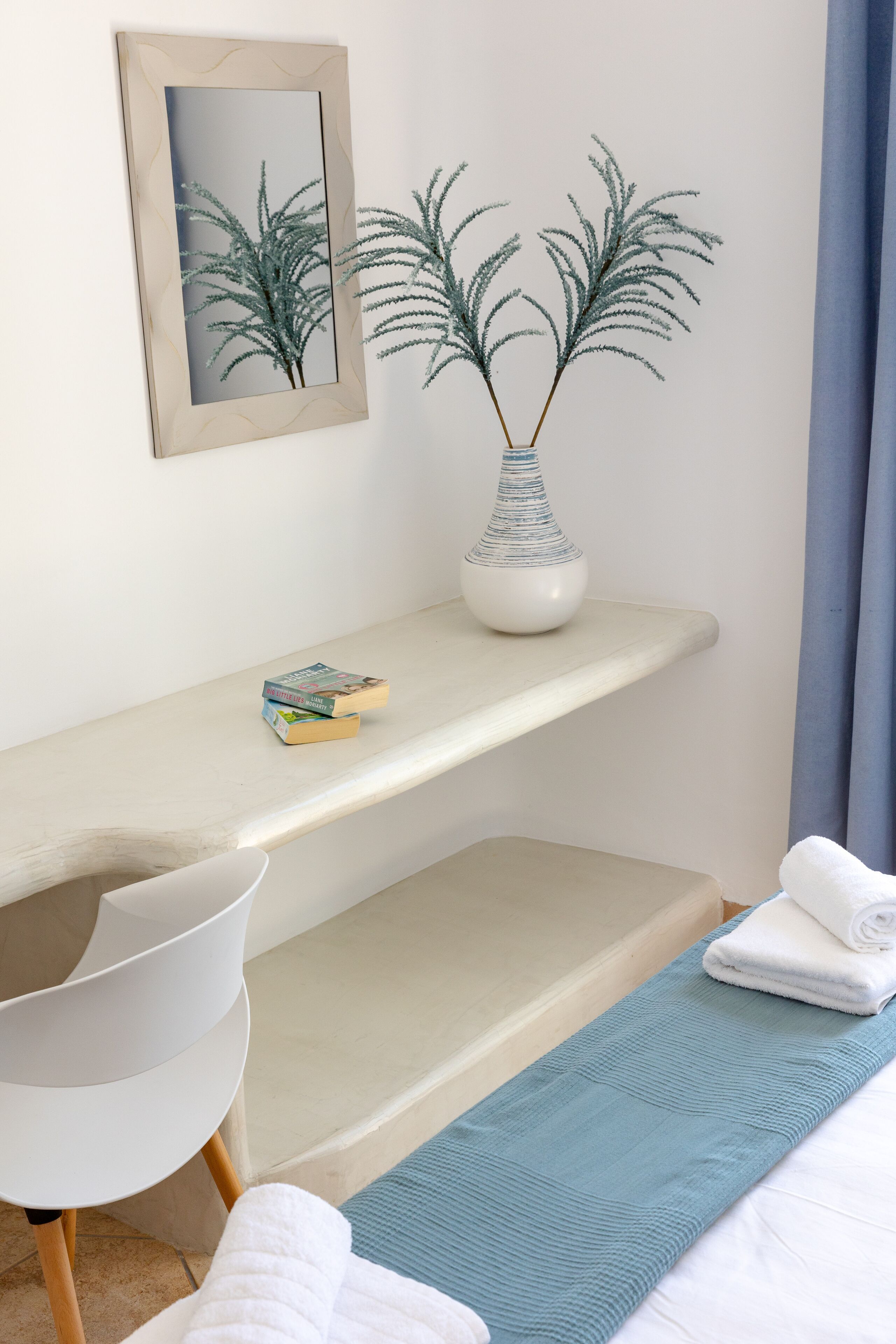 Une chambre d’hôtel bien rangée, avec un lit, une chaise, une petite table sur laquelle se trouvent un vase et des livres, et un tableau encadré au mur.